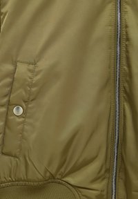 Close-up of olive green jacket fabric with a metal snap pocket, zipped front, and ribbed hem detail.