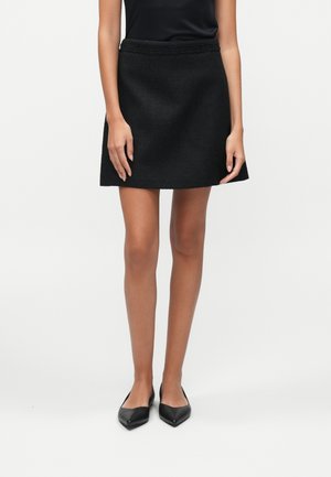 Femme portant une mini-jupe noire texturée, un haut noir sans manches et des ballerines noires à bouts pointus, debout devant un fond uni.