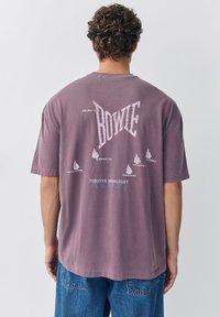 Man met krullend haar die een verweerd paars David Bowie tour t-shirt draagt met op de achterkant de namen van Canadese steden, gecombineerd met een blauwe spijkerbroek.