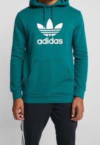 Person som bär en teal Adidas hoodie med vit trefoil-logga och svarta byxor med vita sidosträck, står mot en enfärgad bakgrund.