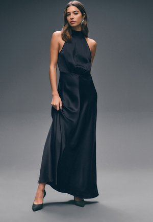 Kobieta w czarnej, bezrękawnikowej sukni z dekoltem halter i czarnych szpilkach, pozująca na szarym tle, lekko trzymająca sukienkę.