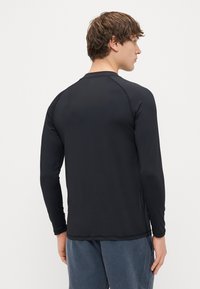 Czarna sportowa koszulka z długim rękawem o gładkiej fakturze, z rękawami raglanowymi i zaokrąglonym dołem, noszona z casualowymi niebieskimi spodniami.