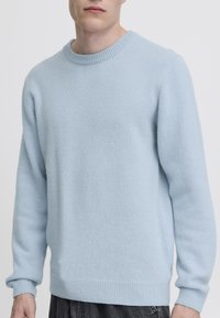 Moški v svetlo modrem puloverju z okroglim izrezom, ribastimi manšetami in robom, stoji pred enobarvnim belim ozadjem.