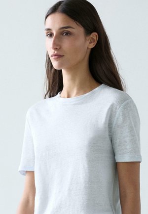 Žena s dlouhými tmavými vlasy, oblečená v jednoduchém bílém tričku s krátkými rukávy a kulatým výstřihem, mírně otočená nalevo na světle šedém pozadí.