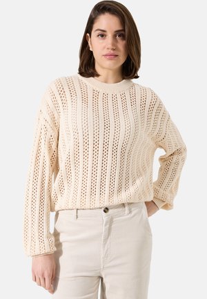 Pull en tricot crème avec motif ajouré, doté d'un col côtelé et de manches longues. Associé à un pantalon en velours côtelé de couleur claire.