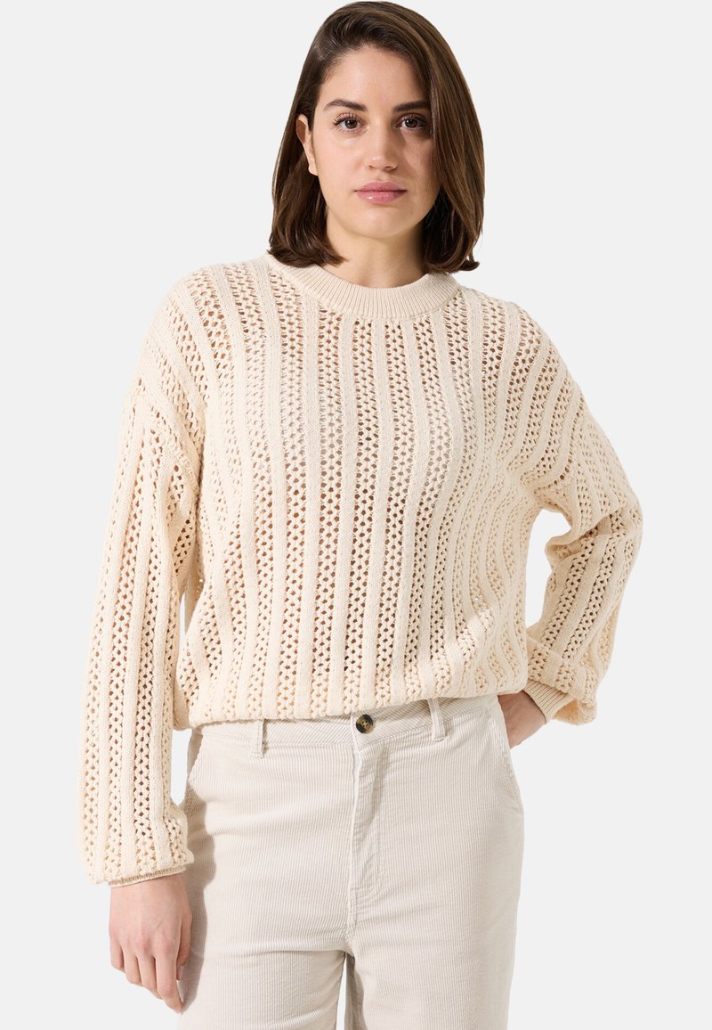 Pull en tricot crème avec motif ajouré, doté d'un col côtelé et de manches longues. Associé à un pantalon en velours côtelé de couleur claire.