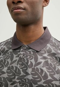 Primer plano de una persona que lleva una camiseta de polo gris oscuro con patrones abstractos de hojas y tres botones en la tapeta.