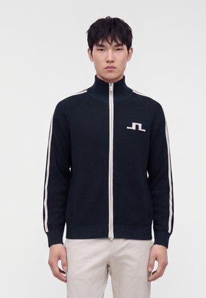 Junger Mann trägt einen marineblauen Pullover mit Reißverschluss, weißen Streifen an den Ärmeln und weißem Logo auf der Brust, kombiniert mit hellen Hosen.