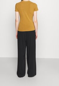 Senapsgul kortärmad topp i kombination med lösa svarta byxor, visad bakifrån mot en vit bakgrund. Tillverkad av bomull, avslappnad passform.