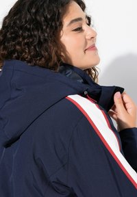 Námořnická modrá bunda s kapucí, bílými a červenými pruhy, hladkou texturou a předním zipem.