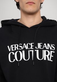Hoodie noir en coton, avec un logo imprimé blanc "VERSACE JEANS COUTURE". Accentué par des cordons de serrage et une poche kangourou.