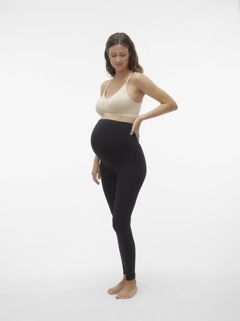 Grūtniece stāv basām kājām, ģērbusies bežā grūtniecības krūšturī un melnā grūtniecības legingos, ar roku uz apakšējā muguras daļas, maigi smaidot.