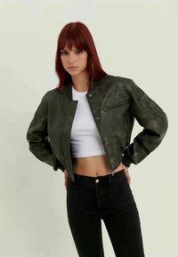 Ung kvinne med rødt hår som har på seg en mørkegrønn bomberjakke, hvit cropped tank tops og svarte høyhårede bukser, står med hendene på hoftene.