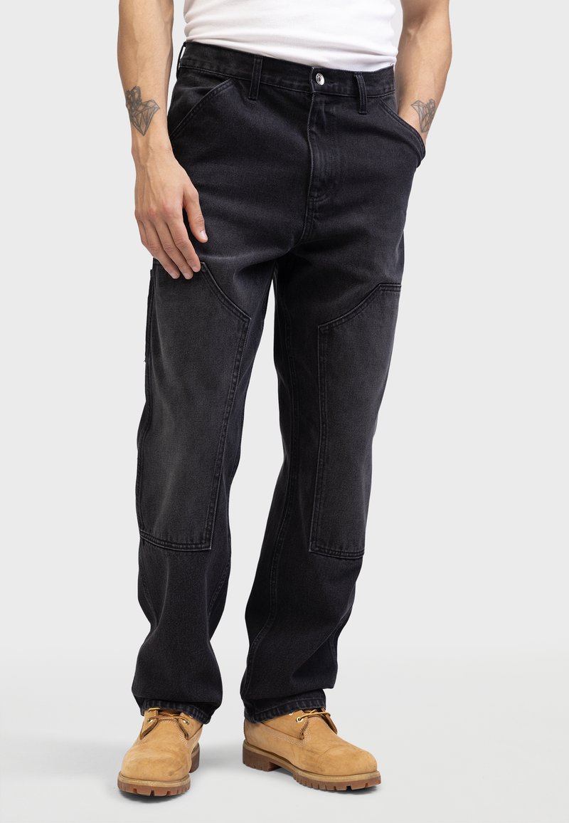 Sort denimbukser med en afslappet pasform, der har sidelommer og unikke lapdetaljer. Båret sammen med beige snørebåndsstøvler.