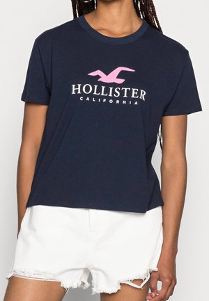 Personne portant un t-shirt bleu marine Hollister California avec un logo oiseau rose associé à un short en denim blanc effiloché.