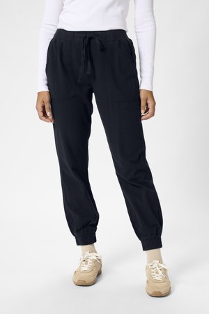 Osoba ubrana w czarne spodnie dresowe ze sznurkiem i elastycznymi mankietami, beżowe trampki oraz białą bluzkę z długim rękawem, stojąca na tle jednolitego tła.