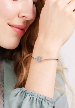 Zilveren armband met een ronde bedel met een uitgesneden ontwerp. Draag op een pols tegen een lichtgroene stoffen achtergrond. Gladde textuur.