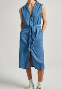 Jeansklänning i ljusblått, ärmlös med krage, framknappar och knytbälte. Mittenlång med en slits framtill för rörelsefrihet.