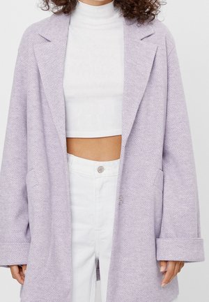 Femme portant un manteau oversize violet clair par-dessus un col roulé court blanc et un pantalon blanc taille haute sur un fond uni.