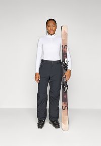 En kvinne står selvsikkert i skiklær, holder ski ved siden av seg. Hun har på seg en tettsittende topp og baggy bukser, klar for snøsportsaktiviteter.