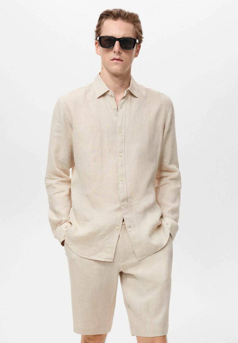 Camisa de lino beige claro con mangas largas, cierre delantero abotonado y pantalones cortos a juego. Llevada con gafas de sol oscuras, textura suave.