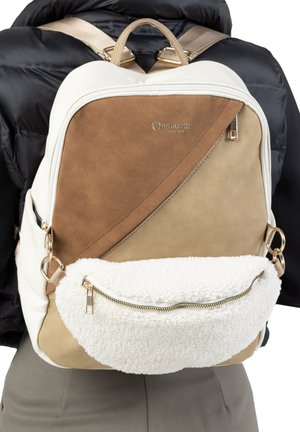 Rucksack mit beigen und braunen Wildlederpaneelen sowie einer vorderen Tasche aus weißem Sherpa-Fleece, getragen über einer schwarzen Jacke und grauen Hosen.