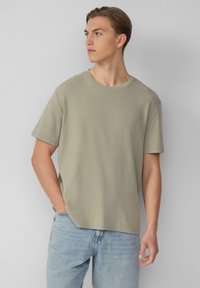 Jasnooliwkowa koszulka z krótkim rękawem i okrągłym dekoltem, o fakturze ściągacza, noszona z jasnoniebieskimi dżinsami.