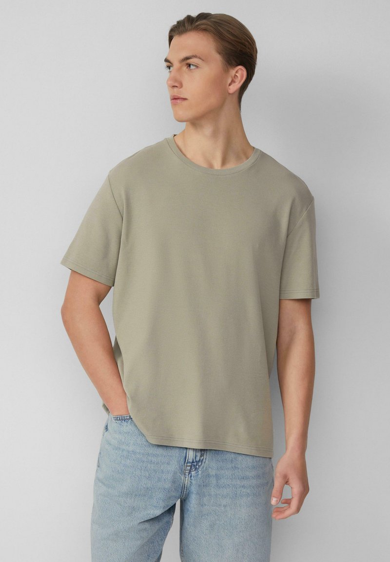 Jasnooliwkowa koszulka z krótkim rękawem i okrągłym dekoltem, o fakturze ściągacza, noszona z jasnoniebieskimi dżinsami.