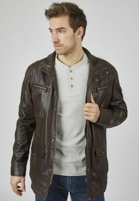 Braune Lederjacke mit Reißverschluss vorne, zwei seitlichen Taschen und einer Reißverschlusstasche auf der Brust. Mit einem Stehkragen und einer leicht abgenutzten Textur.
