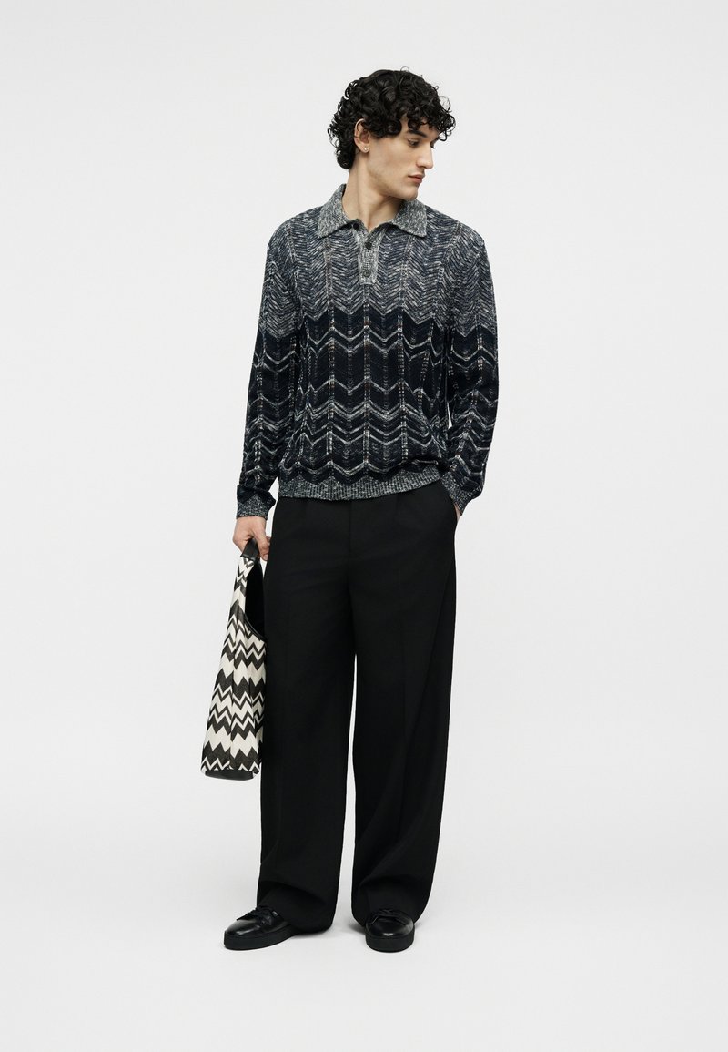 Man with curly hair wearing a patterned long-sleeve knit shirt, wide black pants, and black shoes, holding a black-and-white chevron bag.