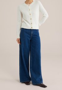 Vit stickad cardigan med rund halsringning och gyllene knappar, i kombination med vida mörkblå jeans och spetsiga svarta skor.