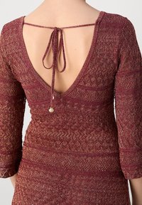 Robe bordeaux avec un dos en V et des manches trois-quarts, ornée de motifs texturés et d'un détail noué au cou avec un accent en forme de coquillage.