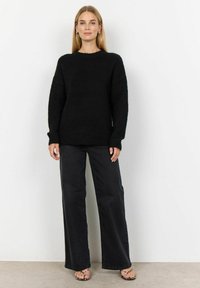 Pulover negru din tricot cu decolteu rotund, croi oversized și mâneci lungi, asortat cu pantaloni negri cu croi larg și sandale cu barete.