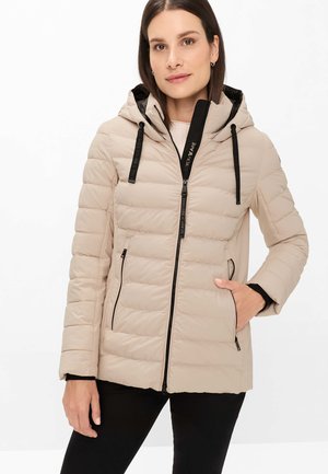 Frau trägt beige gesteppte Jacke mit schwarzem Reißverschluss, Kapuze und Kordeln, schwarze Hose, eine Hand in der Tasche, neutraler Hintergrund.