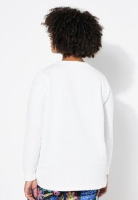 Sudadera blanca con texto en relieve en la parte trasera, cuello redondo y puños acanalados, combinada con pantalones de colores con estampado.