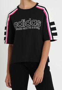 Černé bavlněné tričko s kulatým výstřihem, výrazným bílým a růžovým logem Adidas a třemi pruhy na rukávech v růžových a bílých akcentách.