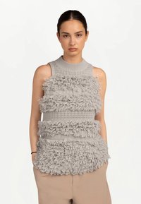 Jeune femme aux cheveux foncés portant un haut gris sans manches en maille avec franges horizontales texturées et un pantalon beige, debout les mains dans les poches.