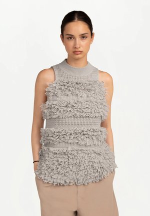 Junge Frau mit dunklen Haaren, die ein ärmelloses graues Stricktop mit texturiertem, horizontalem Fransenmuster und beigen Hosen trägt, steht mit den Händen in den Taschen.