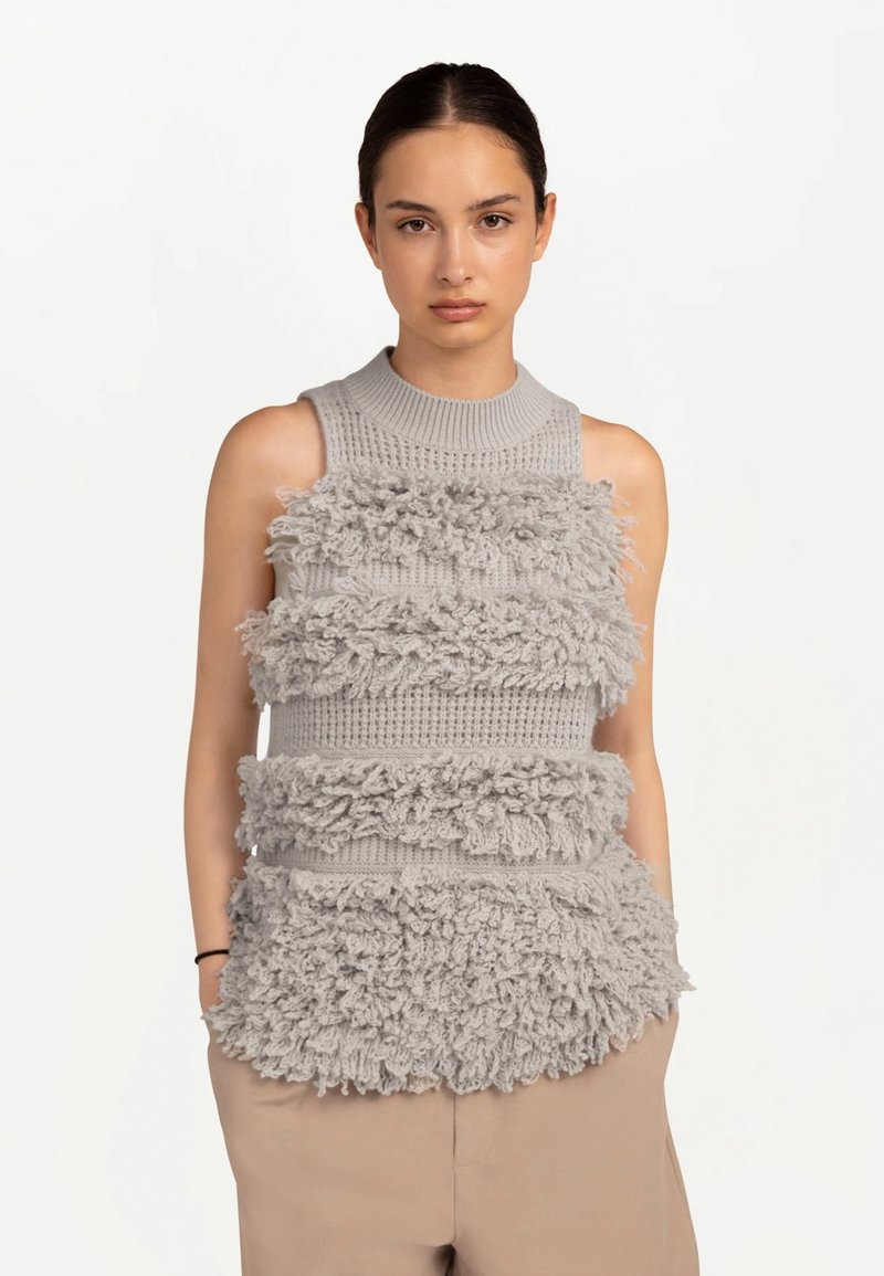 Jeune femme aux cheveux foncés portant un haut gris sans manches en maille avec franges horizontales texturées et un pantalon beige, debout les mains dans les poches.