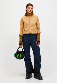 Beige zip-up fleecejakke med hætte, brune detaljer og marineblå snesko. Holder en sort hjelm med grønlig visir. Sorte støvler.