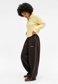 Gul långärmad pikétröja kombinerad med lösa bruna mjukisbyxor med en blå sidrand och logotyp. Outfiten har en avslappnad passform och stil.