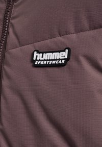 Lila kviltad tyg med en framträdande svart och vit "hummel SPORTSWEAR" logopatch, med en mjuk, texturerad yta.