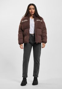 Brun pufferjakke med fleece-overpaneler, broderet "KARL" logo, lynlås lukning og ribbede manchetter. Sammen med mørke jeans og ankelstøvler.