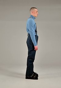 Homme en col roulé bleu, bretelles noires, pantalon de ski noir et chaussures de ski, debout de profil devant un fond uni.