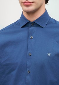 Tamsiai mėlyna mygtukų marškinėliai iš lygaus audinio su apykakle, su subtiliu logotipu išsiuvinėtu ant krūtinės ir suderintais mygtukais.