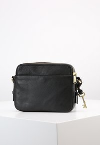Schwarze, strukturierte Leder-Crossbody-Tasche mit goldenem Reißverschluss und angehängtem goldenen Schlüssel-Anhänger, platziert auf einem weißen Sockel vor einem schlichten Hintergrund.