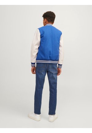 Dreng vendt væk iført blå varsityjakke med hvide ærmer, blå jeans og hvide sneakers, stående på ensfarvet baggrund.