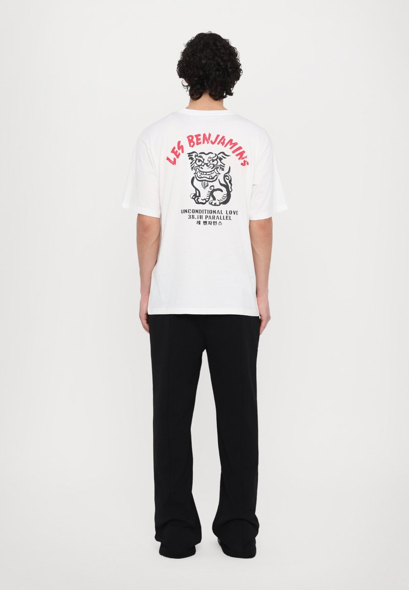 T-shirt blanc à manches courtes avec impression graphique au dos, présentant un design de tigre et du texte rouge. Associé avec un pantalon noir.