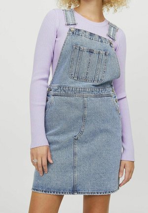 Personne portant un haut à manches longues côtelé violet clair sous une robe salopette en jean bleu avec poche avant et détails à boutons.