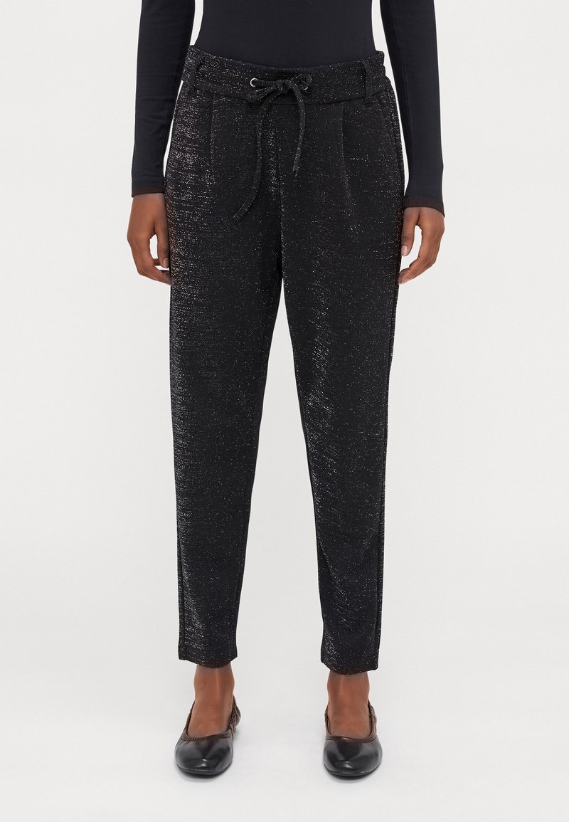 Pantalon noir fuselé avec une texture subtilement brillante, taille à cordon, porté avec un haut noir à manches longues et des chaussures plates noires.